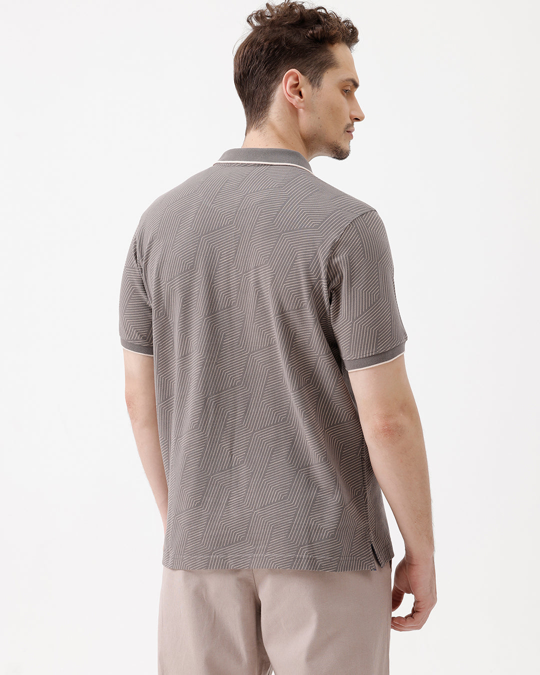 A man wearing a short-sleeve polo shirt with a gray and white geometric pattern, paired with beige trousers, standing against a plain white studio background.