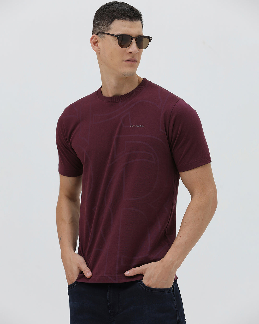 A man wearing a short-sleeve crewneck t-shirt in maroon with a subtle tonal graphic pattern, paired with dark blue jeans, against a light gray studio background.