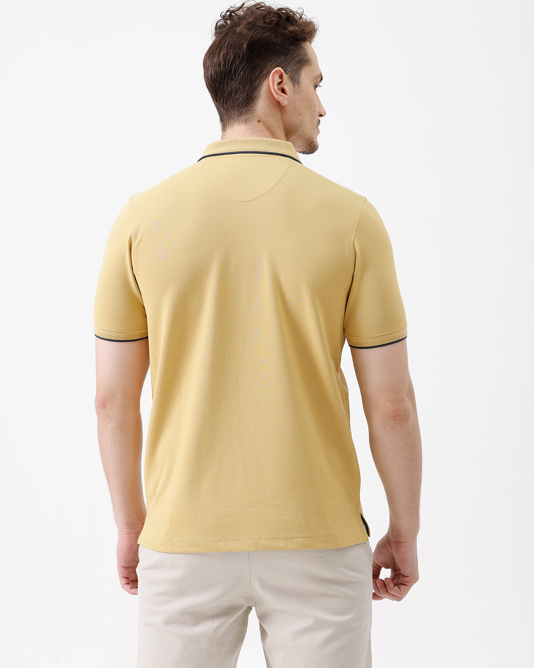 A man wearing a light yellow Lacoste polo shirt with dark piping on the collar and sleeve cuffs, paired with beige trousers, against a plain white studio background.