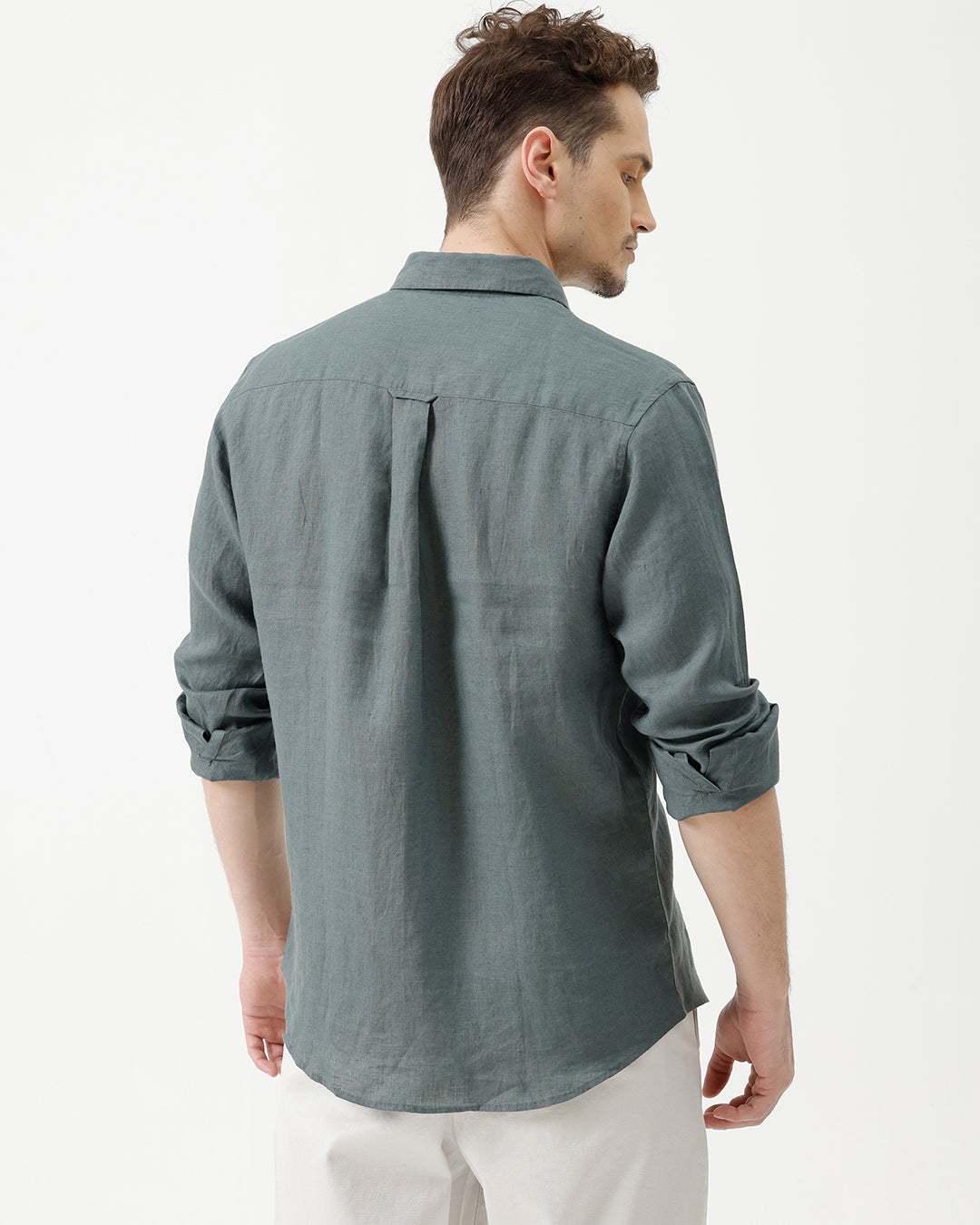 A man wearing a slate blue-green long-sleeve linen button-up shirt with white trousers, standing against a plain white studio background.
