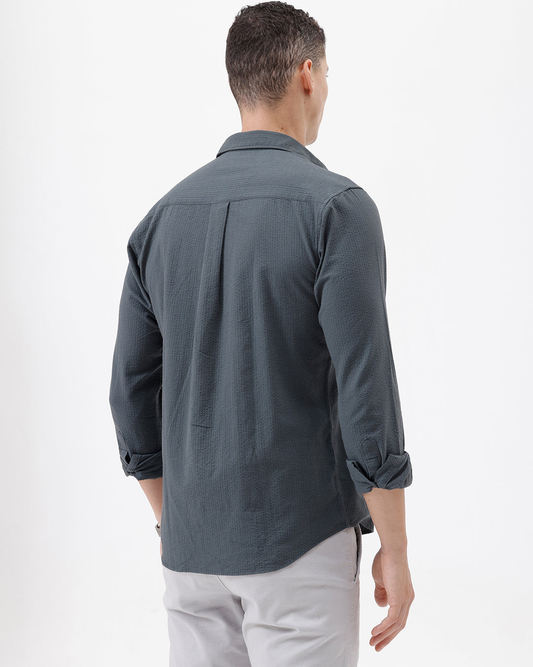 A man wearing a dark charcoal gray long-sleeve button-down shirt with a chest pocket, paired with light gray trousers, standing against a plain white studio background.