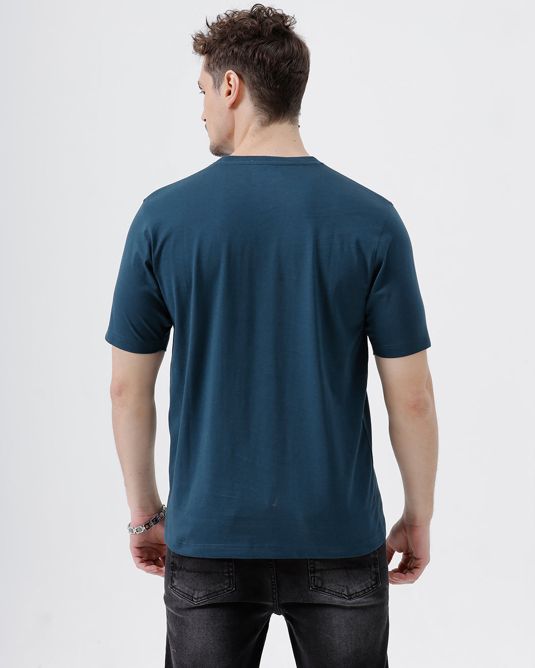 A man wearing a dark navy blue crew-neck t-shirt and black jeans, standing against a light gray studio background.
