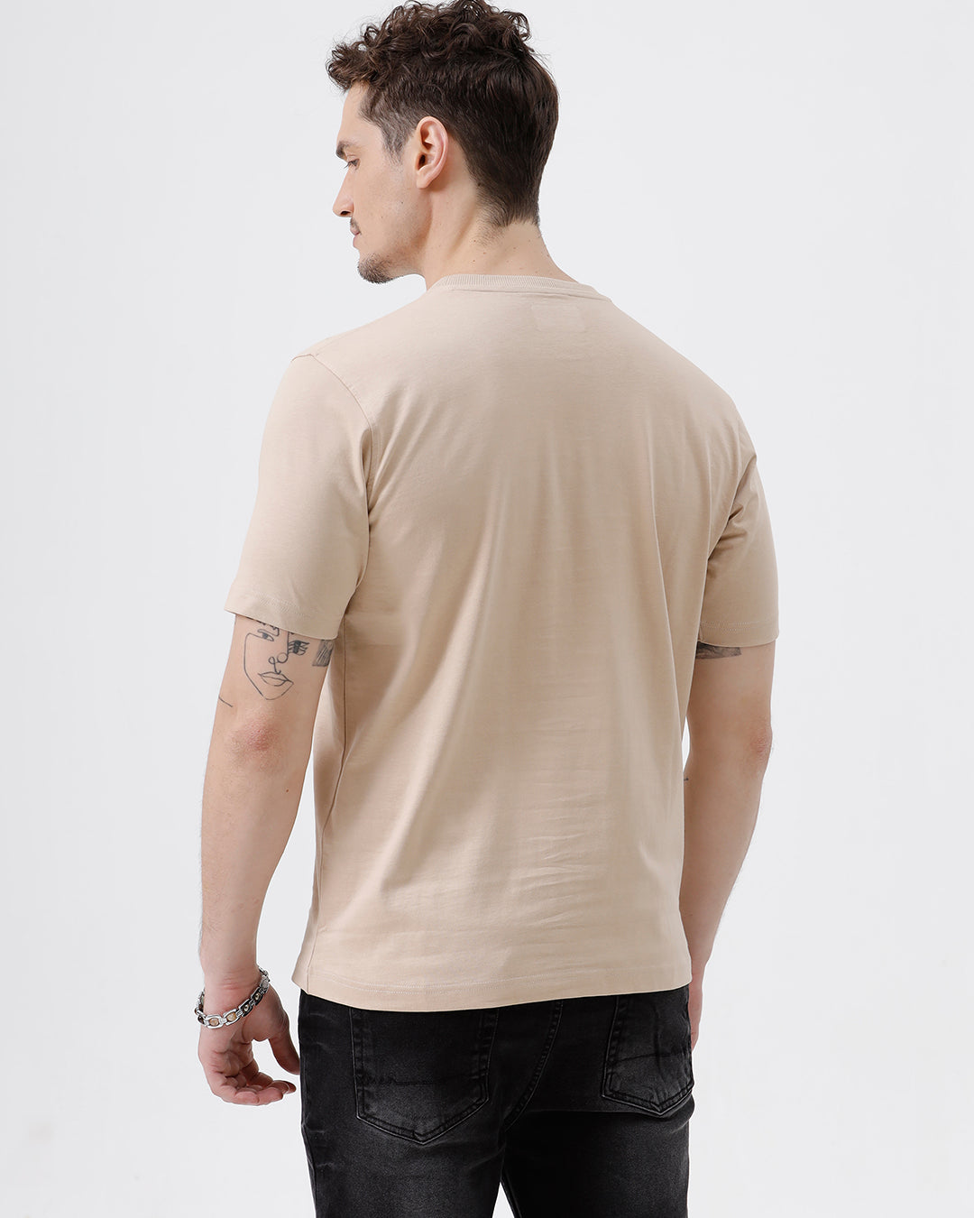 A man wearing a beige short-sleeve crewneck t-shirt with a subtle tonal logo on the left chest, paired with black jeans, standing against a light gray studio background.