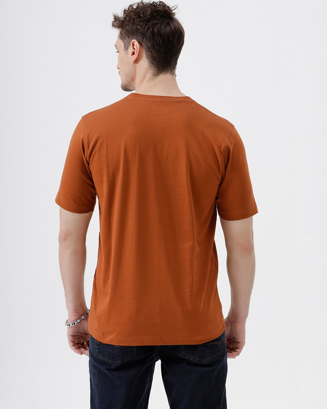 A man wearing a rust-orange crew-neck t-shirt and dark blue jeans, standing against a light gray studio background.