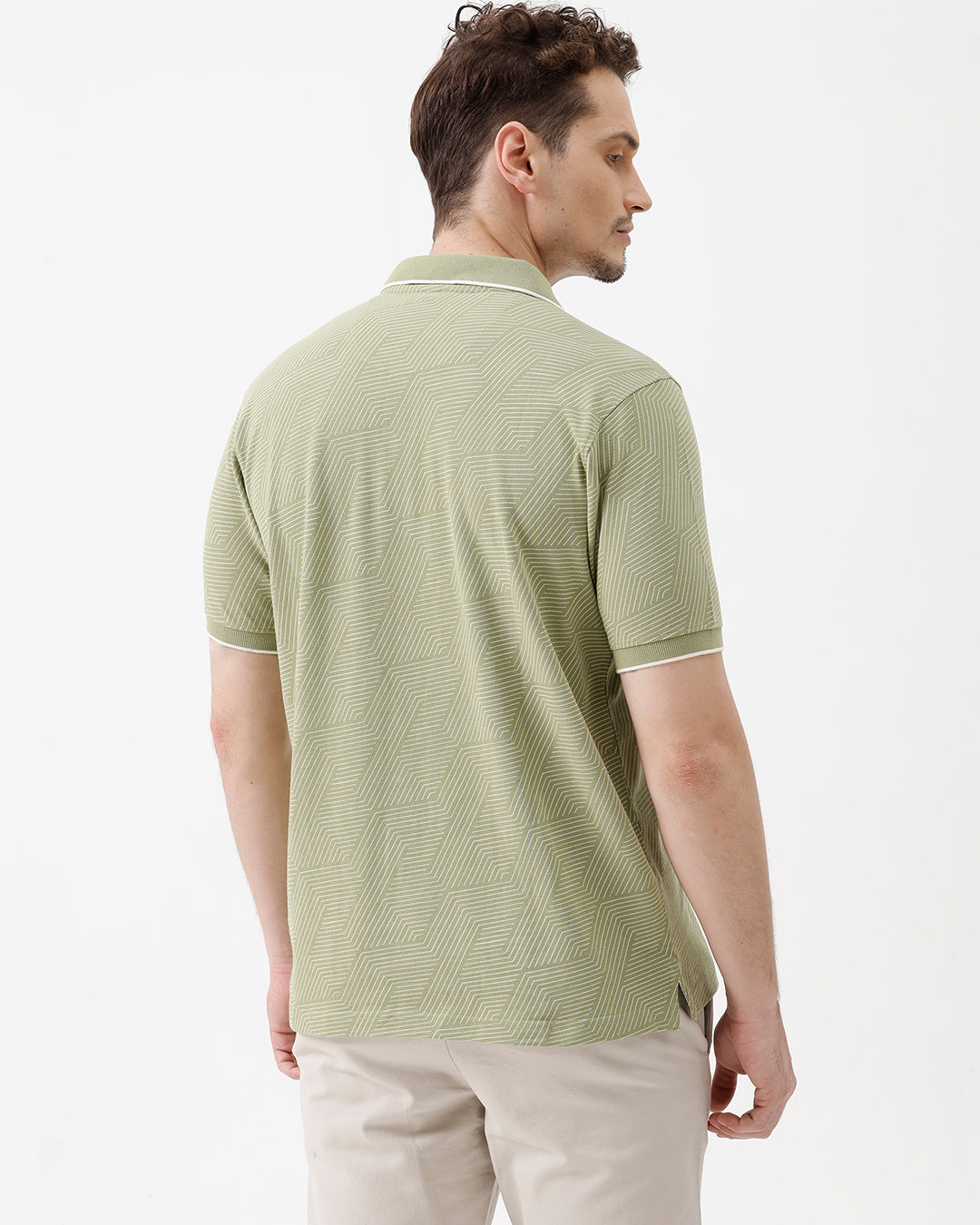 A man wearing a light sage green short-sleeve polo shirt with a subtle geometric pattern, paired with beige trousers, standing against a plain white studio background.