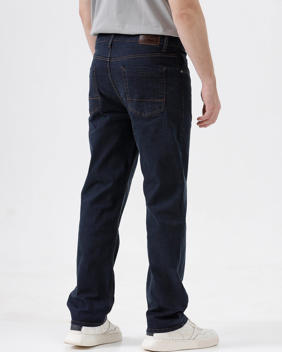 A person wearing dark navy blue straight-leg jeans, a light gray t-shirt, and white sneakers, standing against a plain white studio background.