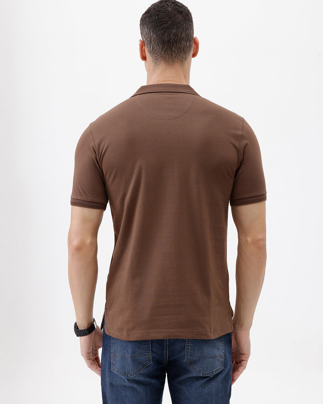 A man wearing a brown short-sleeve polo shirt with a chest pocket, paired with blue jeans, standing against a plain light gray background.