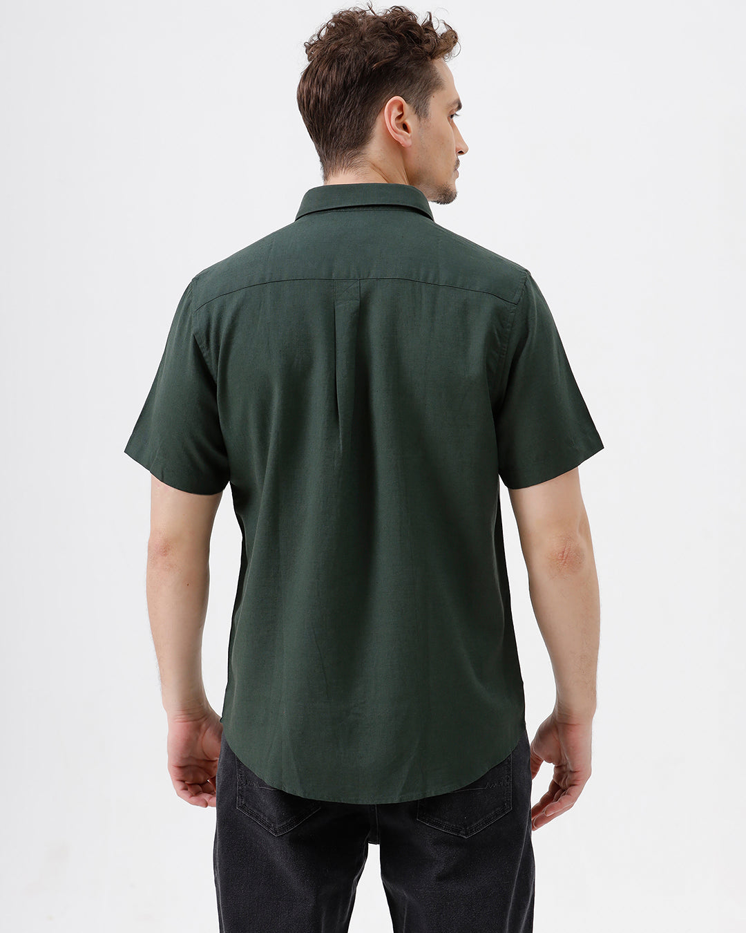 A man wearing a dark forest green short-sleeve button-up shirt with a chest pocket, paired with black jeans, standing against a plain white studio background.