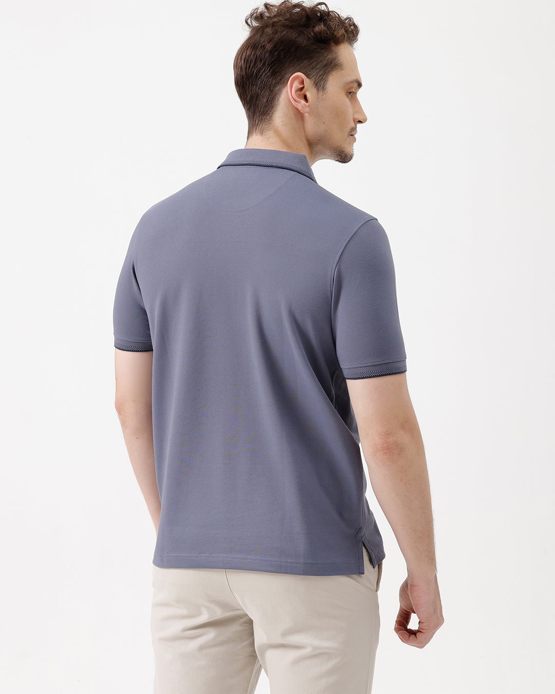 A man wearing a slate blue polo shirt and beige trousers, standing against a plain white background.