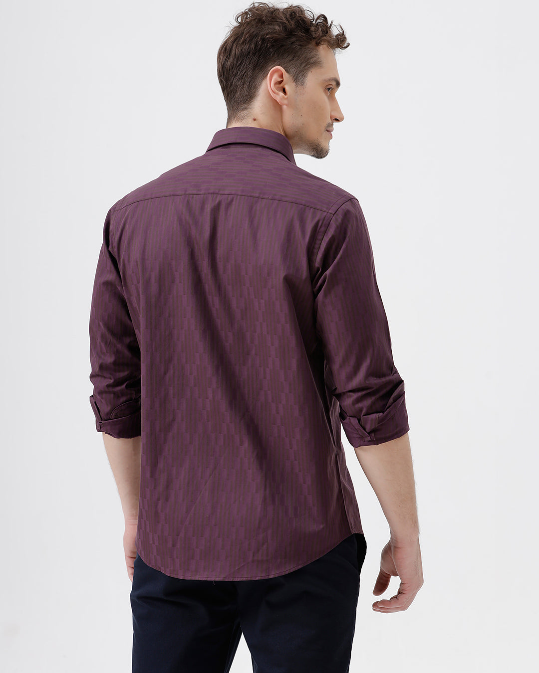 A man wearing a dark purple long-sleeve button-down shirt with subtle vertical stripes and dark navy trousers, standing against a light gray background.