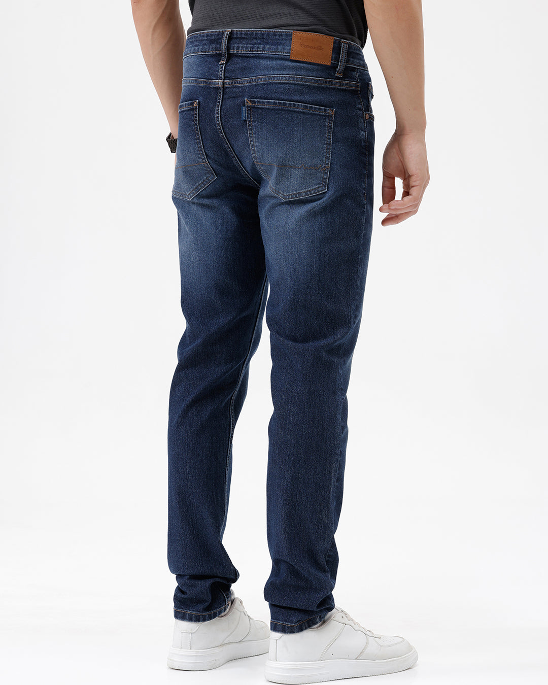 A person wearing dark blue slim-fit jeans with a faded wash, paired with a black t-shirt and white sneakers, shown from the waist down against a plain white studio background.