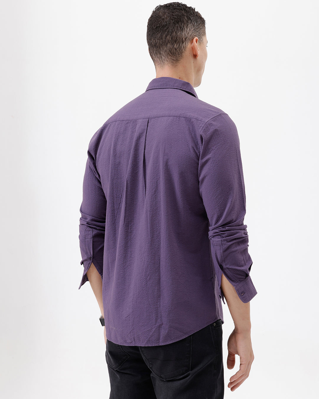 A man wearing a long-sleeve button-down shirt in a muted purple color, paired with black pants, standing against a plain white studio background.