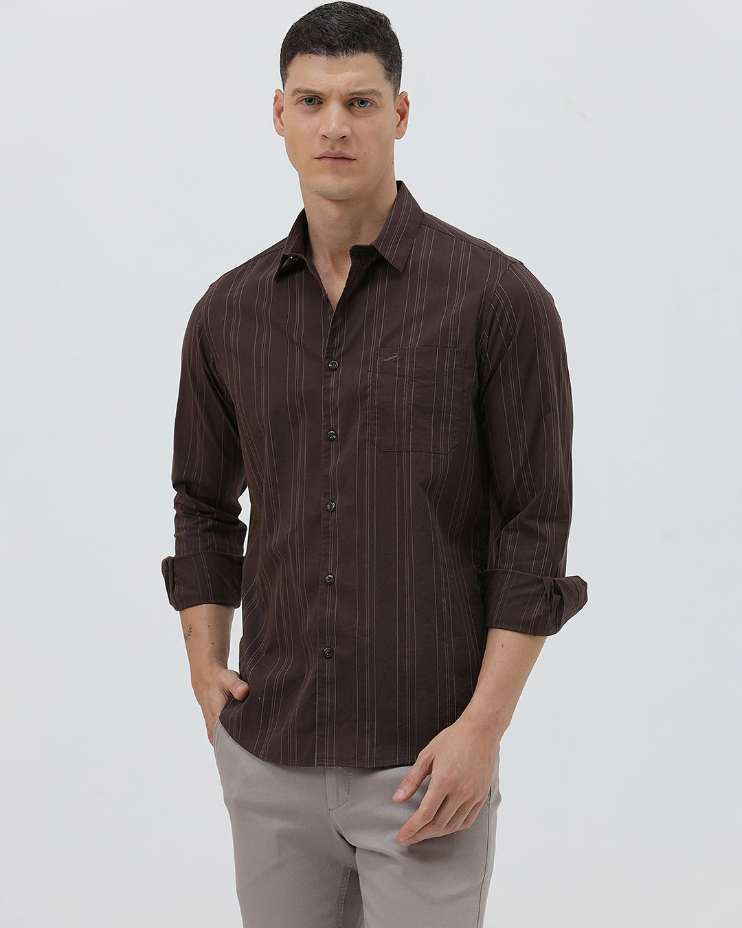A man wearing a dark brown long-sleeve button-up shirt with thin vertical stripes and light gray trousers, standing against a plain light gray background.