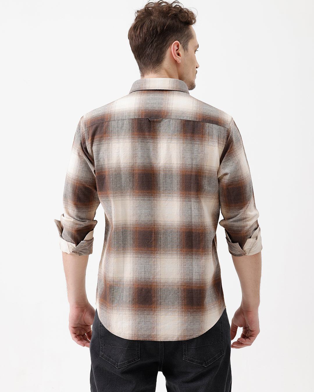 A man wearing a long-sleeve button-up plaid shirt in brown, beige, and gray tones, paired with dark jeans, standing against a plain white studio background.