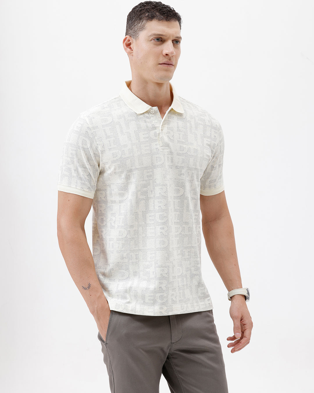 A man wearing a short-sleeve polo shirt with a subtle repeating monogram pattern in cream and light gray, paired with gray trousers, photographed against a plain white studio background.