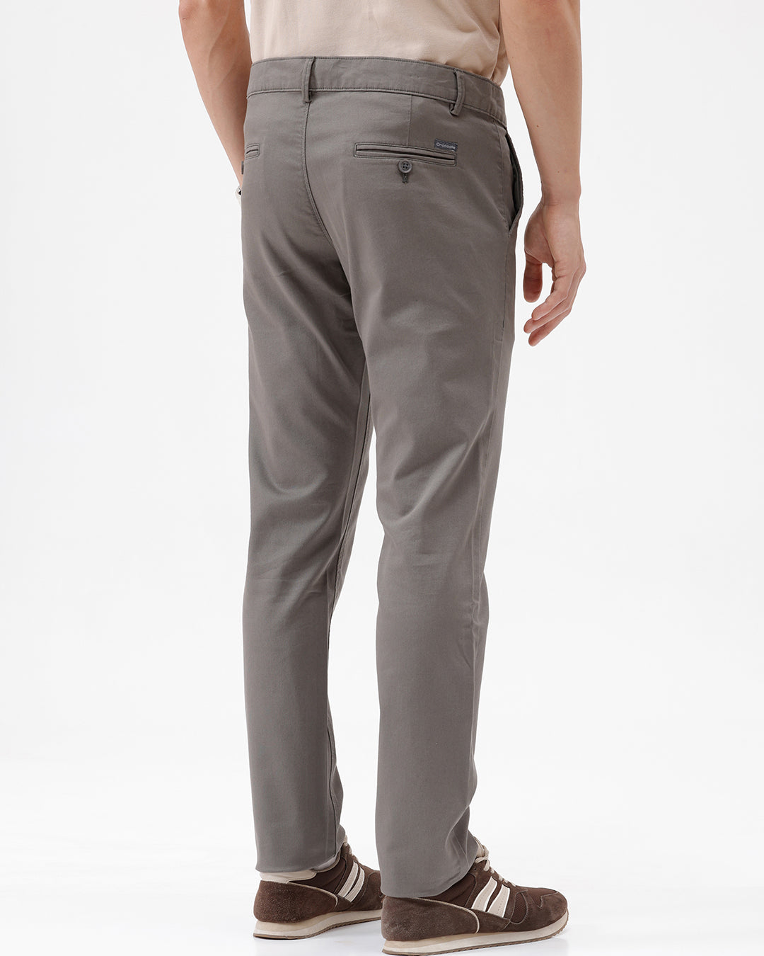 A man wearing slim-fit taupe gray chino pants, paired with a beige t-shirt and brown sneakers, shown from the waist down on a white studio background.