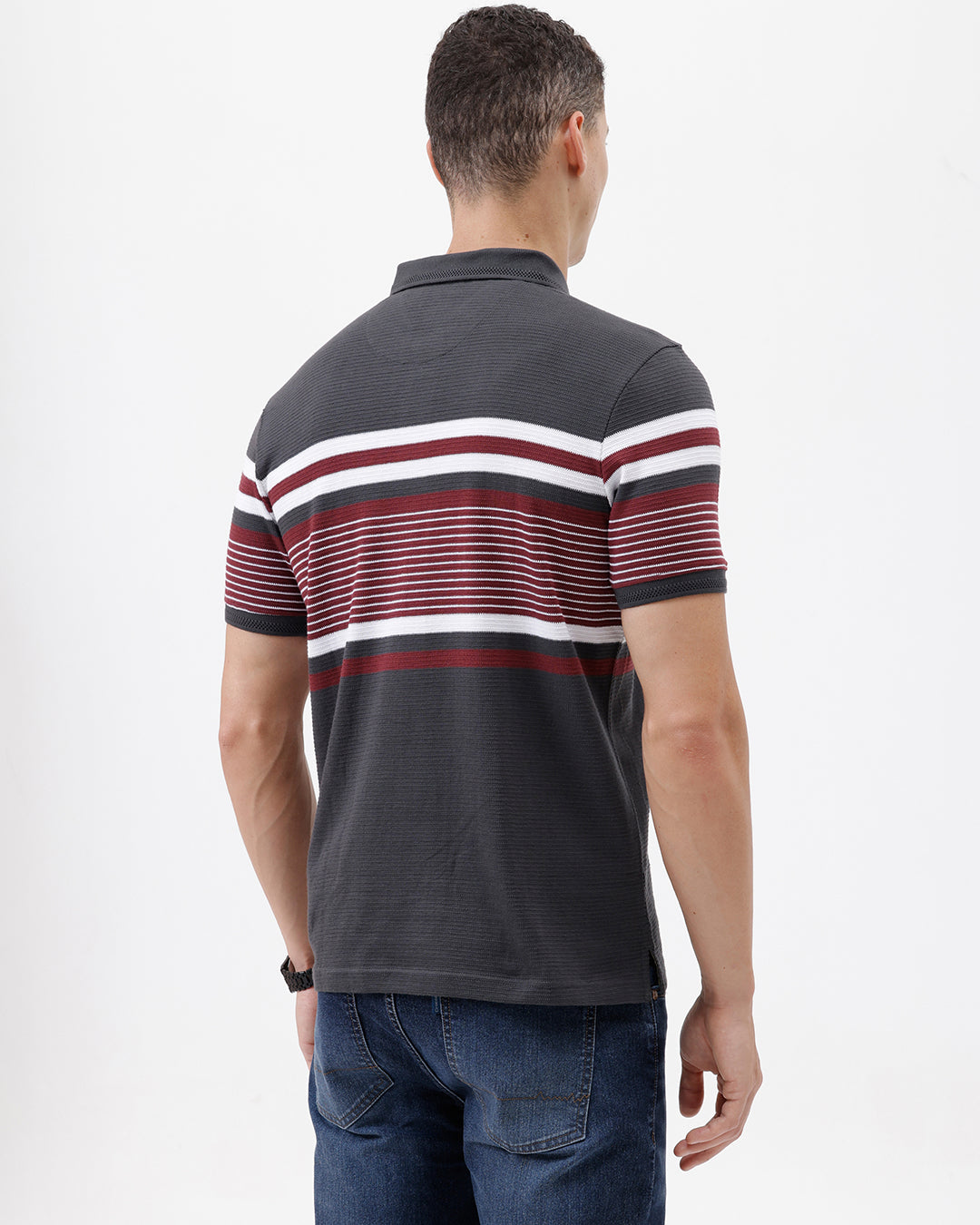 A man wearing a dark gray Lacoste polo shirt with horizontal red and white stripes across the chest and sleeves, paired with blue jeans, against a plain light gray background.