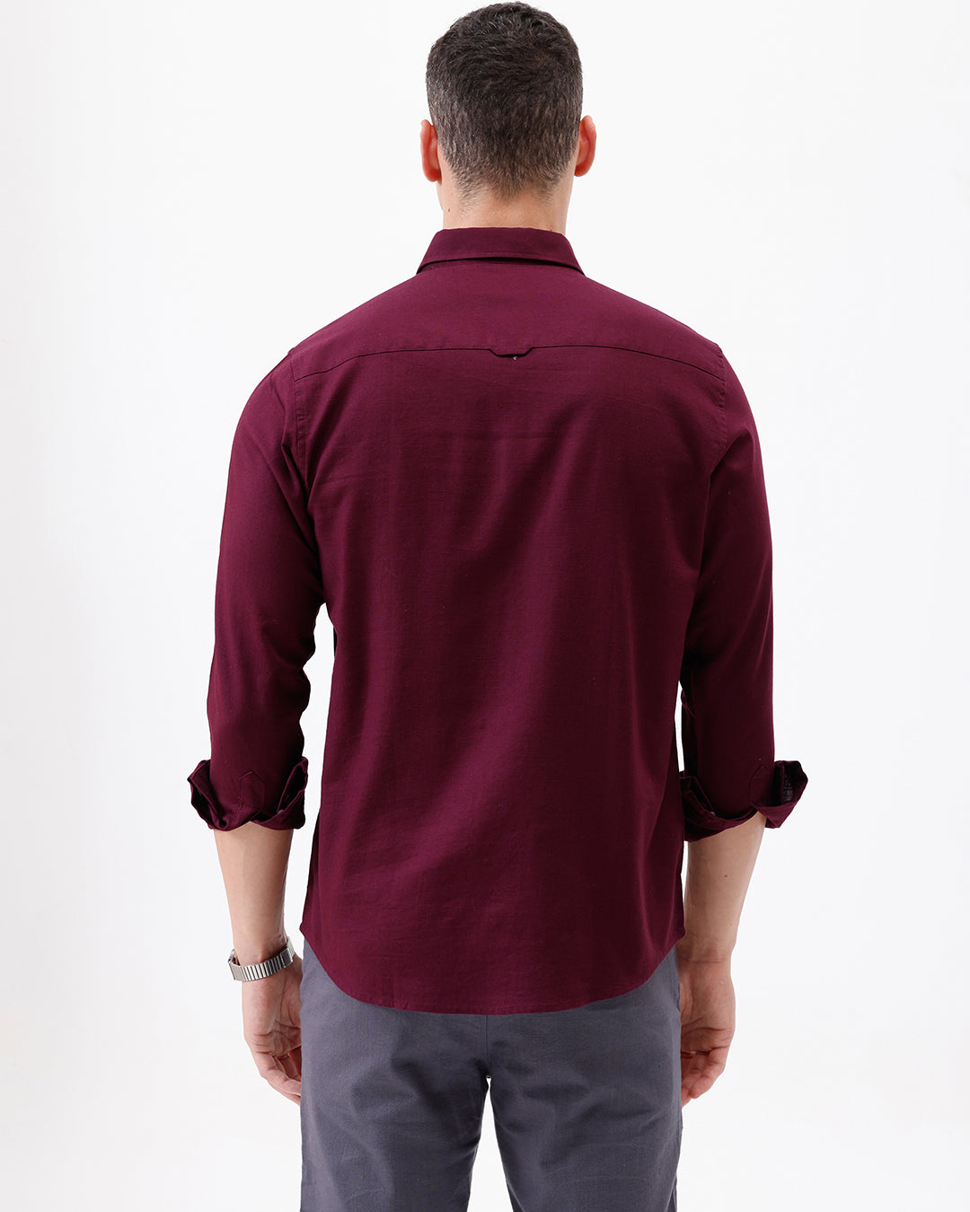 A man wearing a long-sleeve button-down shirt in deep burgundy with rolled-up sleeves and gray trousers, standing against a plain white studio background.