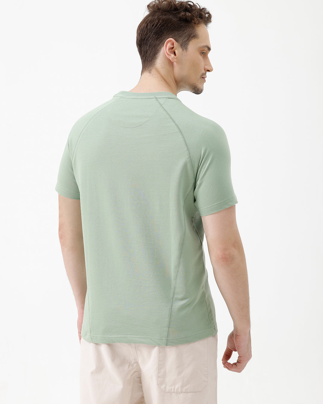 A man wearing a light sage green athletic t-shirt with a small tonal logo on the chest, paired with beige pants, standing against a plain white studio background.