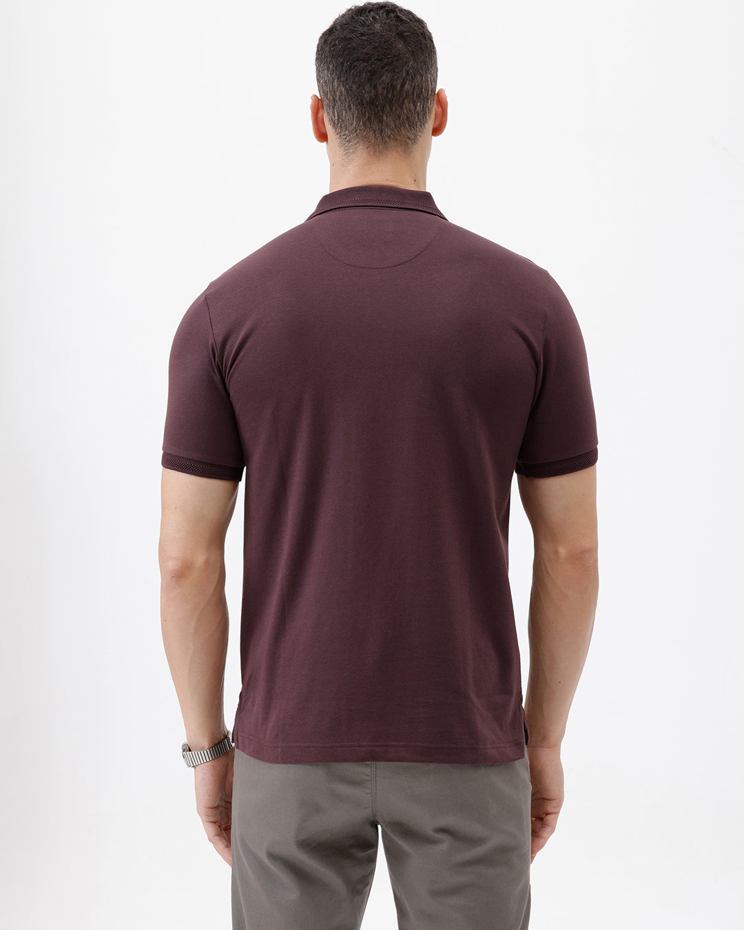 A man wearing a short-sleeve polo shirt in dark burgundy with a chest pocket, paired with gray trousers, standing against a plain light gray background.