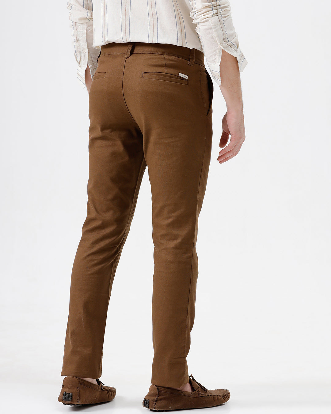 A man wearing brown slim-fit trousers, a cream and brown striped long-sleeve shirt, and brown suede loafers, standing against a plain white studio background.