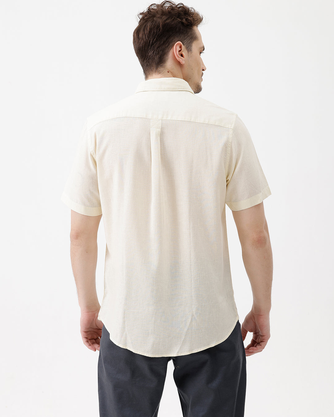 A man wearing a light cream-colored short-sleeve linen button-up shirt with a chest pocket, paired with dark trousers, against a plain white studio background.
