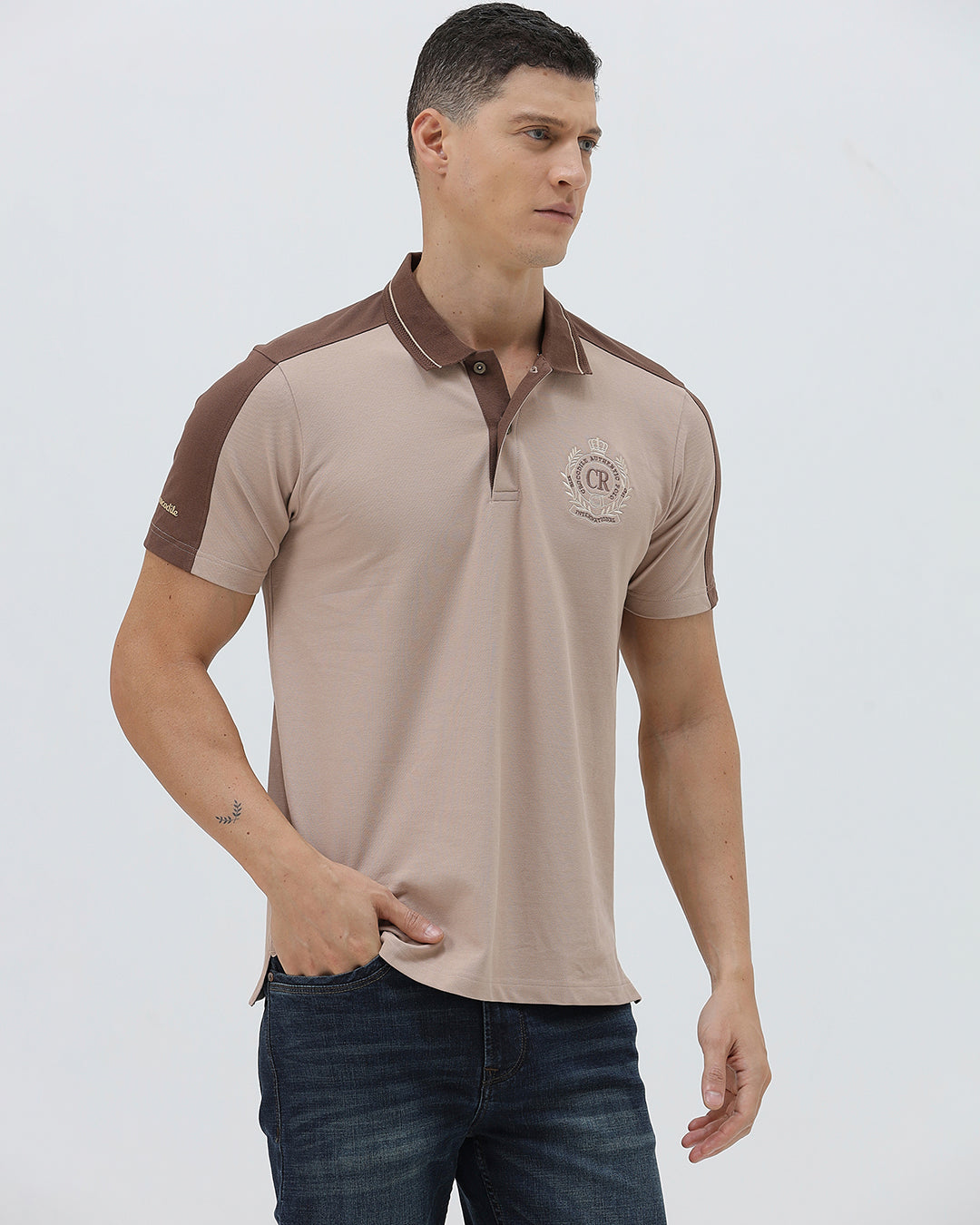A man modeling a short-sleeve polo shirt in beige with brown shoulder panels and collar trim, paired with dark blue jeans, against a light gray studio background.