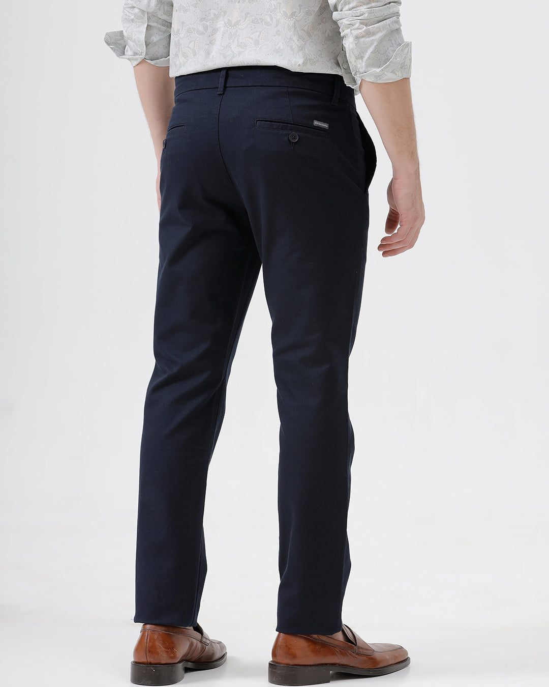 A man wearing dark navy blue straight-leg trousers, a light-colored patterned shirt, and brown leather loafers, standing against a plain white studio background.