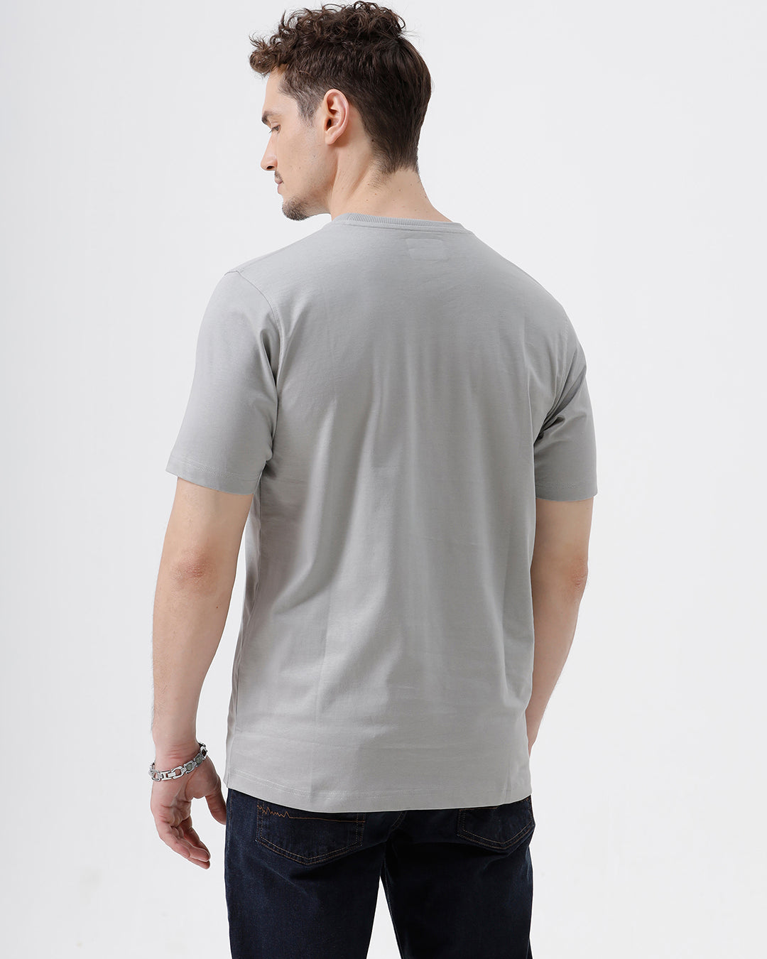 A man wearing a light gray short-sleeve crewneck t-shirt with a subtle tonal chest logo, paired with dark jeans, standing against a light gray studio background.