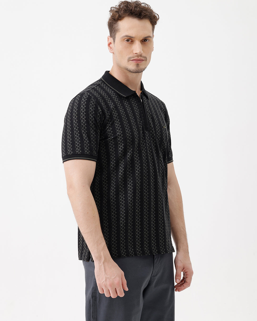 A man wearing a black short-sleeve polo shirt with a subtle geometric pattern, paired with dark gray trousers, standing against a plain white studio background.
