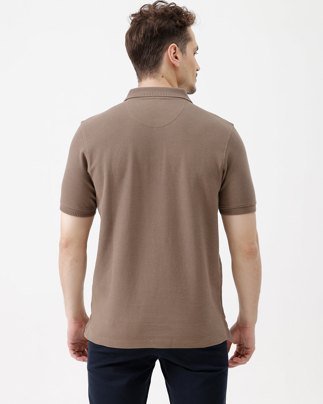 A man wearing a brown short-sleeve polo shirt with dark navy pants, standing against a plain light gray studio background.