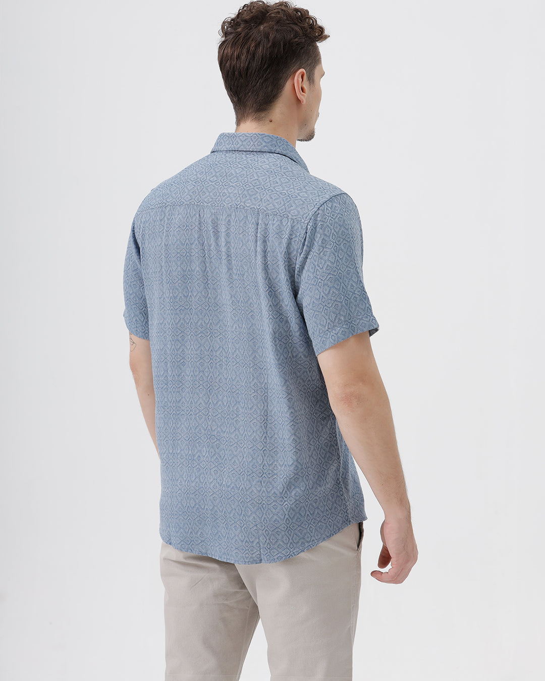 A man wearing a short-sleeve button-up shirt with a fine blue and white geometric pattern, paired with light beige trousers, standing against a plain light gray background.