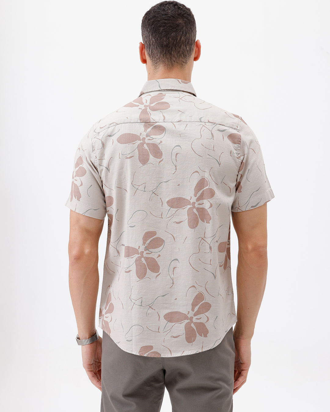 A man wearing a short-sleeve button-up shirt with a beige and brown floral pattern, paired with gray trousers, standing against a plain white background.