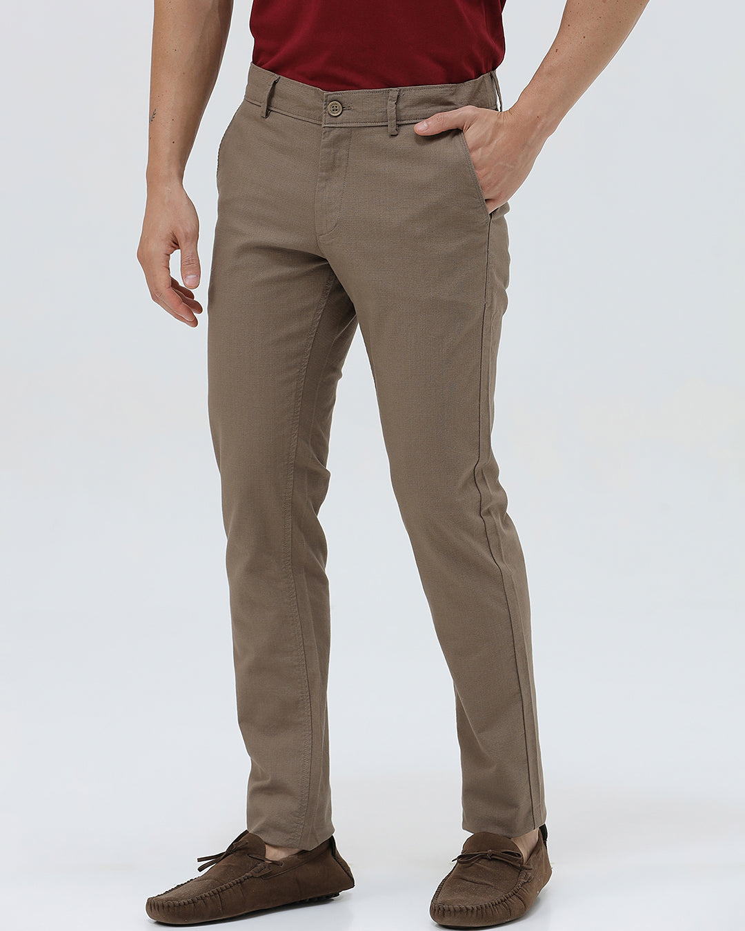 A person wearing slim-fit brown trousers, paired with a dark red shirt and brown suede moccasin shoes, shown from the waist down against a plain white studio background.