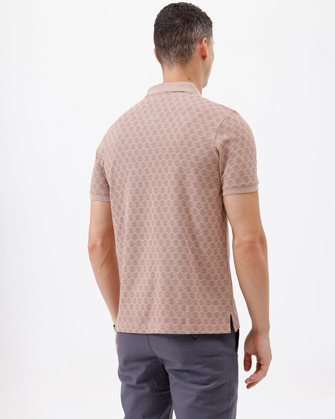 A man wearing a short-sleeve polo shirt with a muted brown and beige geometric pattern, paired with dark gray trousers, standing against a plain white studio background.