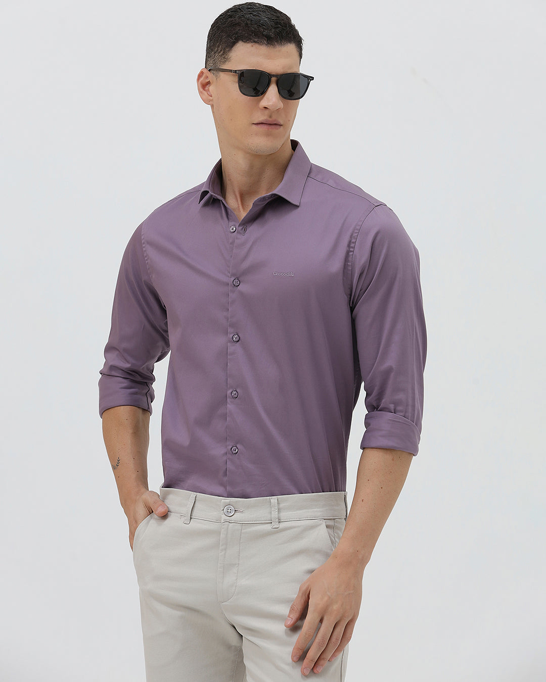 A man wearing a purple long-sleeve button-down shirt with rolled-up sleeves and light beige trousers, standing against a plain light gray background.