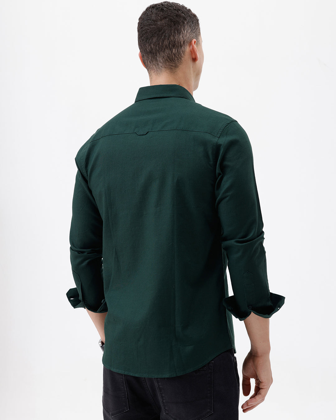 A man wearing a dark forest green long-sleeve button-down shirt with a chest pocket, paired with dark jeans, standing against a plain white studio background.