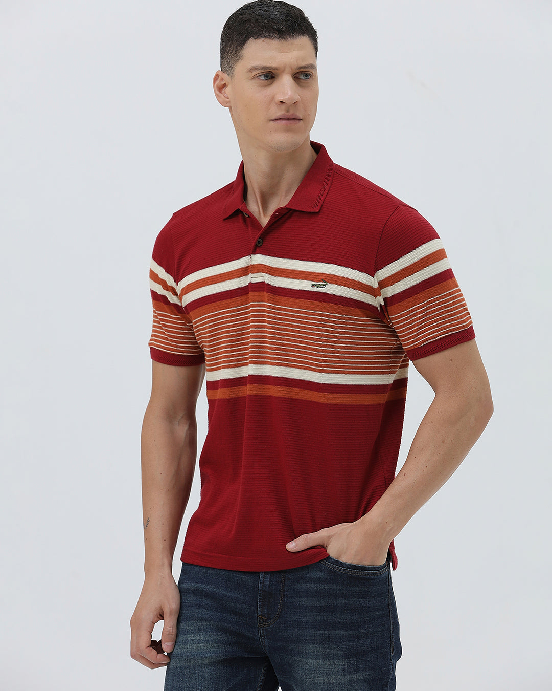 A man wearing a red short-sleeve polo shirt with horizontal cream and rust-orange stripes across the chest and sleeves, paired with dark blue jeans and black sunglasses, against a light gray studio background.
