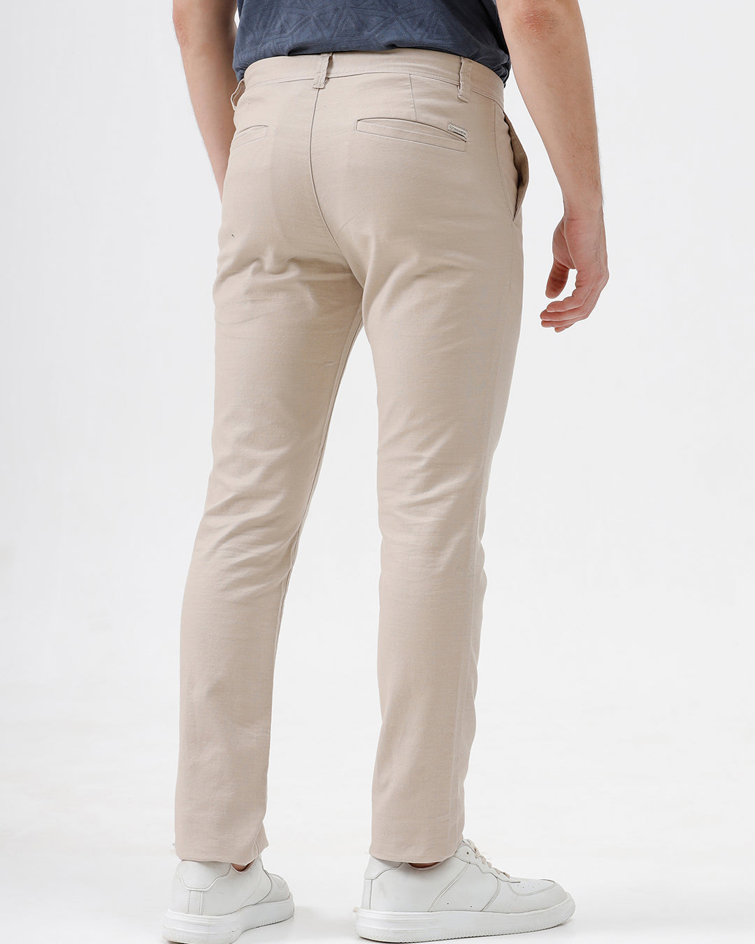 A person wearing slim-fit beige chinos with a blue patterned t-shirt and white sneakers, standing against a white studio background.