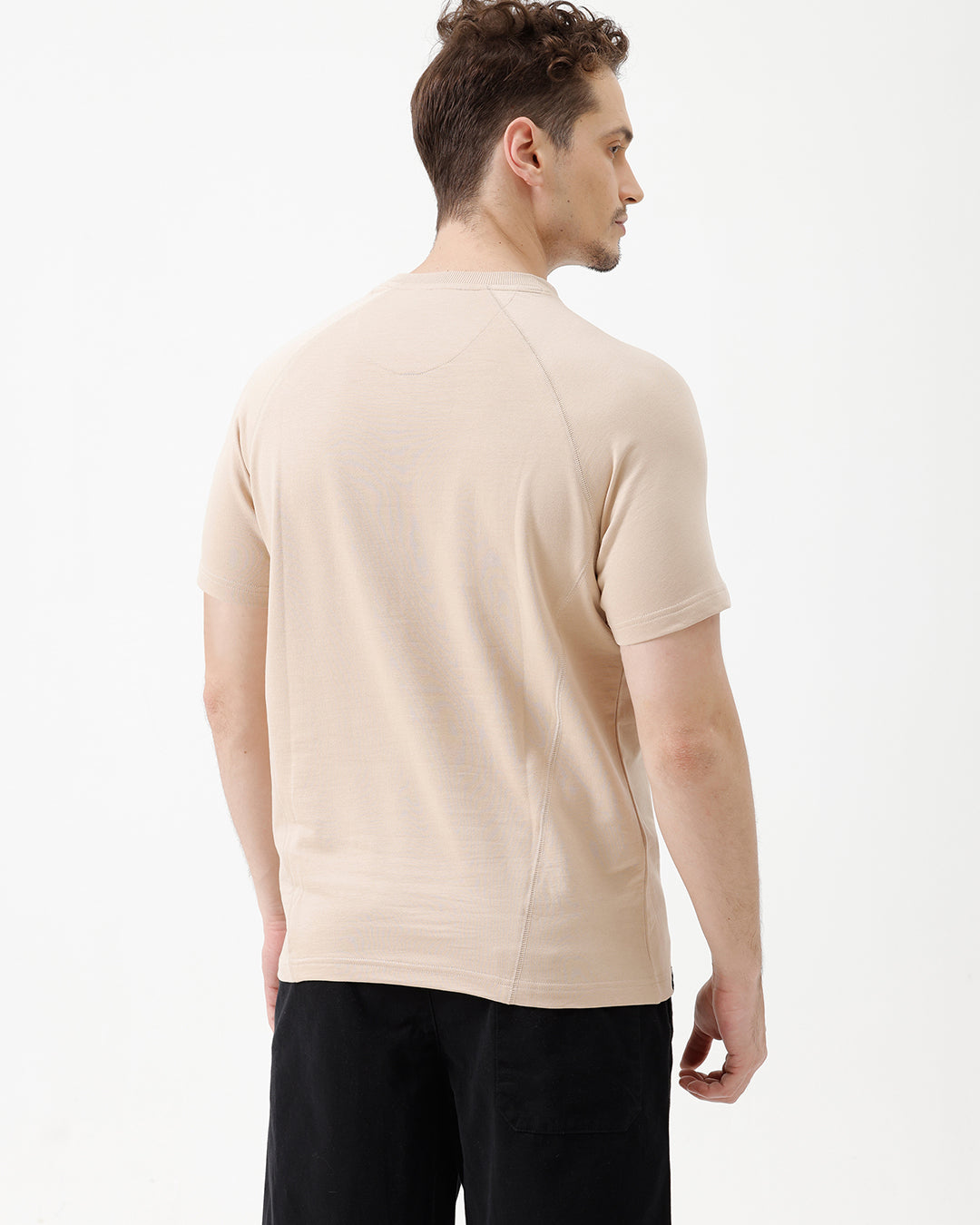 A man wearing a light beige short-sleeve t-shirt with a subtle tonal logo on the chest, paired with black pants, standing against a plain white studio background.