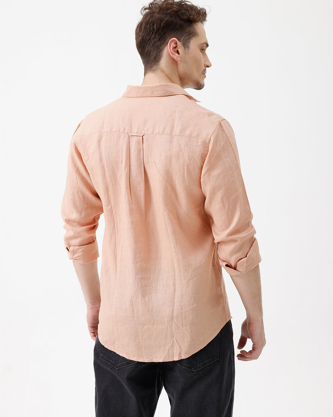 A man wearing a light peach-colored linen button-down shirt with dark jeans, standing against a plain white studio background.