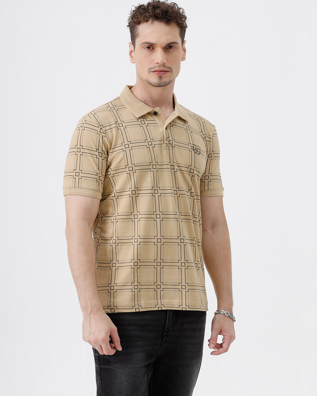 A man wearing a beige short-sleeve polo shirt with a brown grid pattern, paired with black jeans, standing against a plain white studio background.