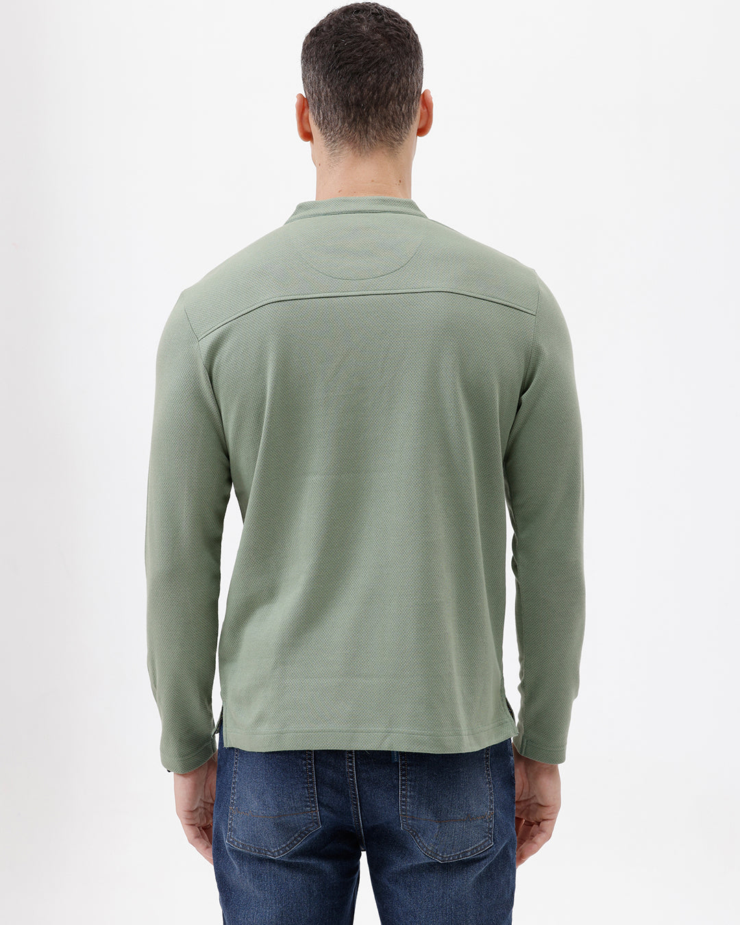 A man wearing a sage green long-sleeve henley shirt with a small embroidered logo on the left chest, paired with blue jeans, against a plain white studio background.