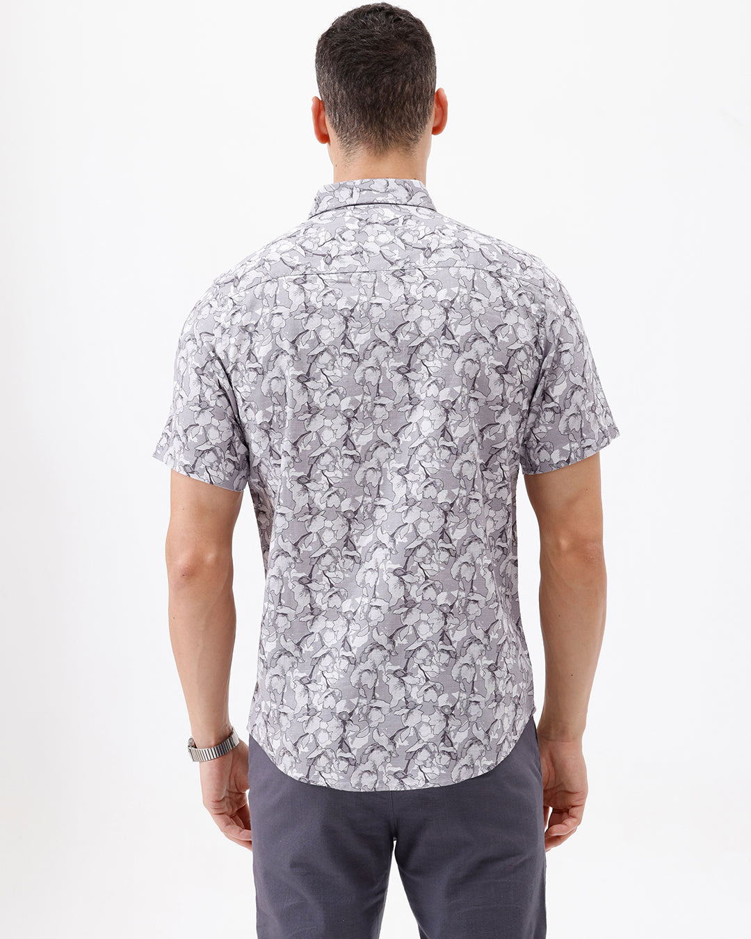 A man wearing a short-sleeve button-up shirt with a gray and white abstract pattern, paired with dark gray trousers, standing against a plain white studio background.