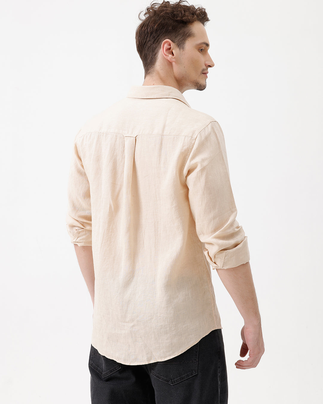 A man wearing a light beige linen button-up shirt with rolled-up sleeves and dark jeans, standing against a plain white studio background.