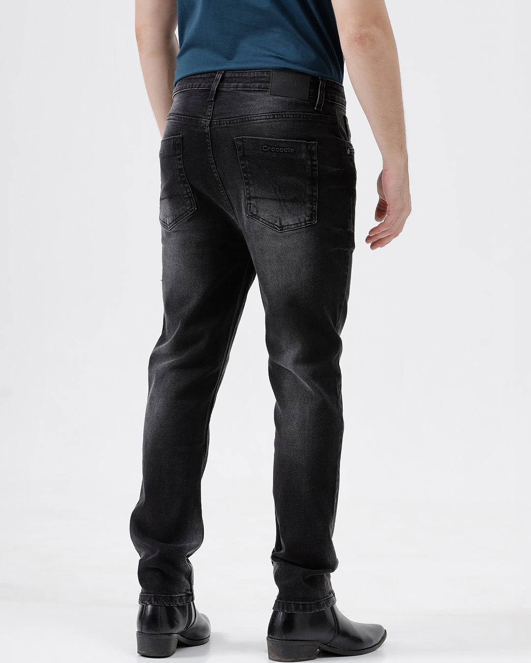 A person wearing slim-fit black denim jeans with a faded wash, paired with a dark blue t-shirt and black leather boots, shown from the waist down on a light gray studio background.