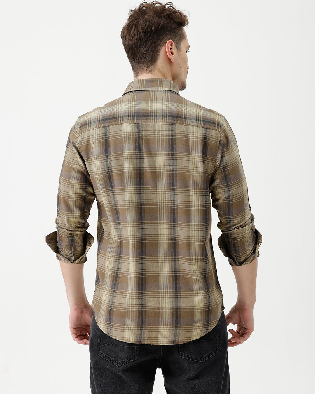 A man wearing a brown and beige plaid long-sleeve button-down shirt with dark sunglasses and black jeans, standing against a plain white studio background.