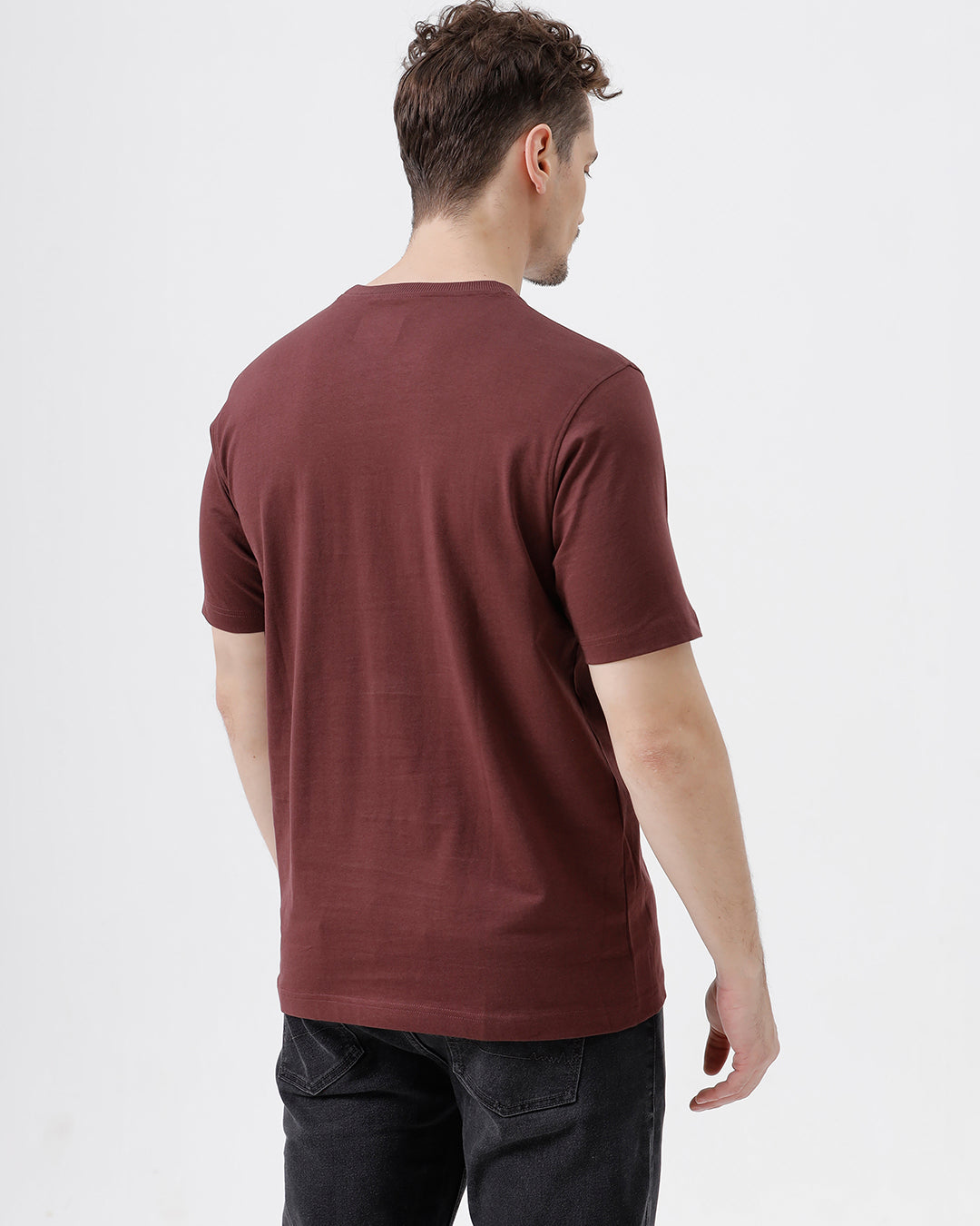 A man wearing a short-sleeve crewneck t-shirt in maroon, paired with dark jeans, standing against a plain light gray studio background.