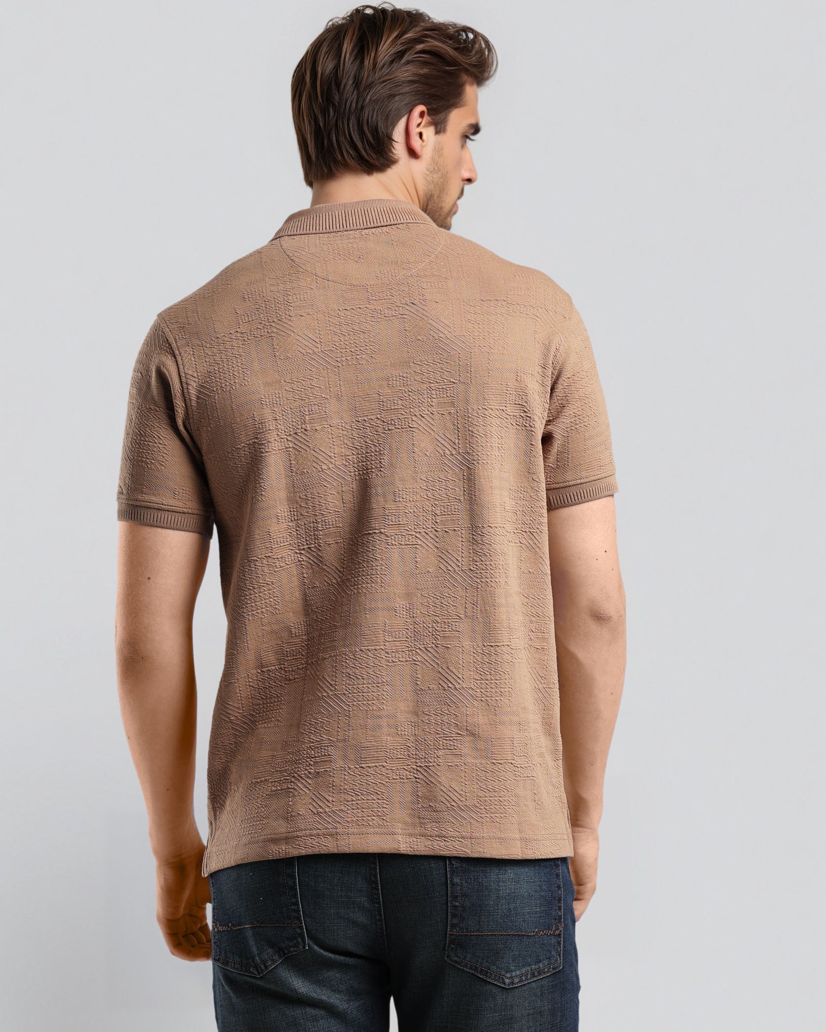 Man wearing a brown patterned polo shirt and dark jeans on a light gray background