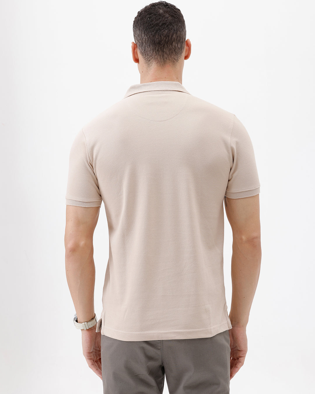 A man wearing a light beige short-sleeve polo shirt with a small crocodile logo on the chest pocket, paired with gray trousers, photographed against a plain white studio background.